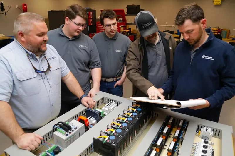 Five team members reviewing a drawing over two control panel backplates being built in the shop