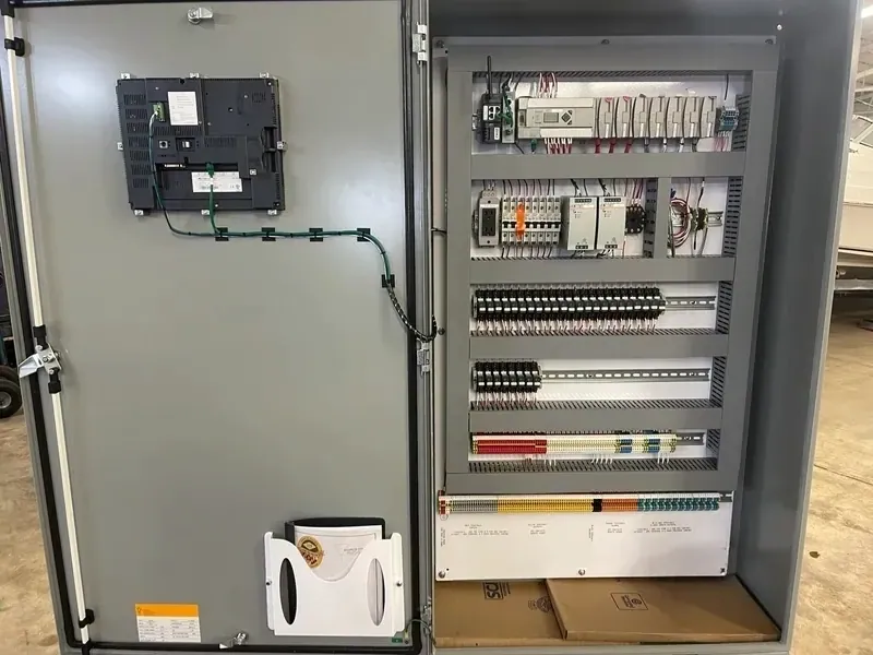 A finished control panel open in the shop: an operator touchscreen on the door, and inside, the PLC, breakers, power supplies, relays and color-coded terminal strips in rows between gray wire duct